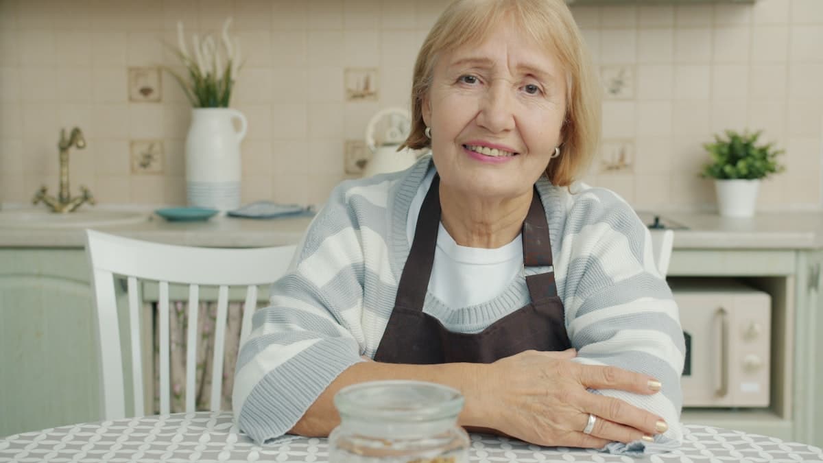 Femme âgée souriante assise dans sa cuisine, attablée devant un repas